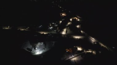 Küçük bir kasabanın üzerinde gece yavaş bir insansız hava aracı uçuşu aydınlanmış sokakları, evleri ve sakin kış atmosferini gösteriyor. Parlayan ışıkları olan huzurlu bir şehir manzarası..