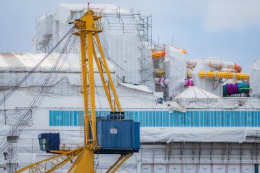 Denize yakın bir tersanede inşa edilen büyük bir geminin fotoğrafı, uzak bir noktadan çekildi. Vinçleri, iskeleyi, büyük gemi yapısını ve endüstriyel deniz ortamını gösterir.