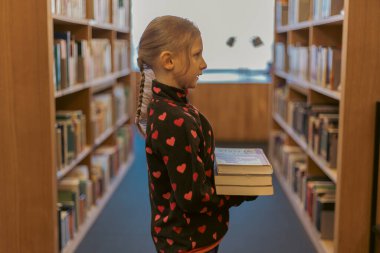 Dişi bir kız kütüphanede elinde bir kitaplık ve pencerenin yanında durup gülümsüyor. Eğitim, okuma, merak, çocukluk, öğrenme, iç mekan, doğal ışık