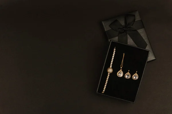İçinde altın bilezikler, uygun küpeler ve siyah lüks hediye kutusunda sunulan zarif bir mücevher seti. Koyu dramatik arkaplan promosyon metni veya sutyeni için mükemmel kopya alanı olan sofistike bir atmosfer yaratır