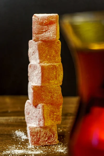 Bir yığın parlak pembe Rahat Lokum, pudra şekerle kaplı, geleneksel Türk çayının yanında duruyor. Cam odağını kaybetmiş ve kısmen kırpılmış dekoratif bir element. Ahşap bir arka planda koyu tonda çekilmiş..