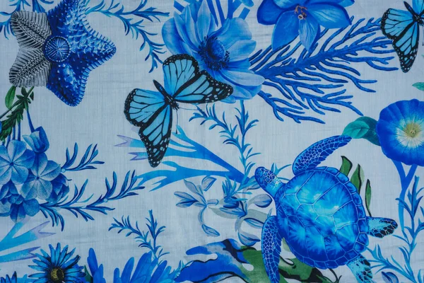 Deniz kaplumbağası, denizyıldızı, kelebek ve canlı mavi tonlarda çiçeklerden oluşan detaylı bir deniz motifi içeren pürüzsüz bir ipek tekstil yüzeyinin yakın çekimi. Tatil yeri giyimi ya da lüks moda için arka plan