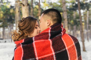 Gelin ve damat, kırmızı bir battaniyeye sarılıp, kış ormanında öpüşüyorlar.