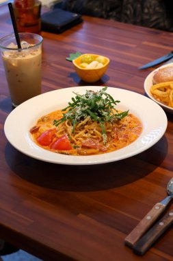 Domates soslu kremalı makarna, taze roka ve parmesan peyniri ahşap masada buzlu kahveyle servis ediliyor. İtalyan mutfağı ve günlük yemek konsepti