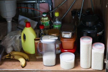 Mutfak lavabosunun altında çeşitli yiyecek kapları, pirinç kavanozları ve ev malzemeleri var. Günlük organizasyonu, bakkaliyeyi, temizlik ürünlerini ve ev içi depolama alanını gösteren gerçek bir ev.