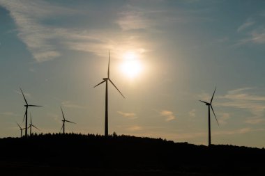 Silhouette of wind turbines at sunset with warm sunlight and soft clouds in the sky over a rural landscape.