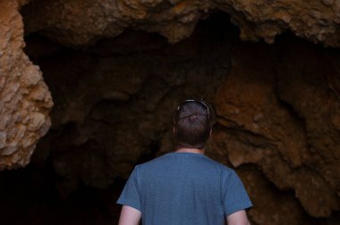 Karanlık kayalık bir mağaraya bakan bir adamın arka görünüşü, keşfi, merakı ve bilinmeyenin gizemini sembolize eder. Doğal kaya dokusuna sahip kavramsal açık hava fotoğrafı..