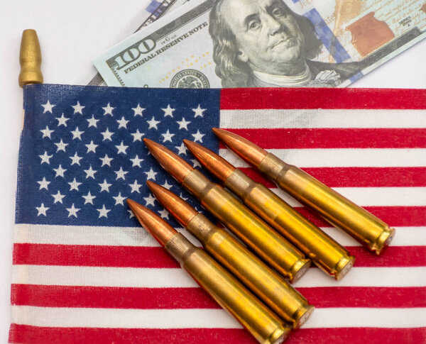 Several rifle bullets placed on a small United States flag with hundred dollar banknotes, symbolizing power, finance, and conflict.