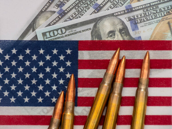 Several rifle bullets placed on a small United States flag with hundred dollar banknotes, symbolizing power, finance, and conflict.