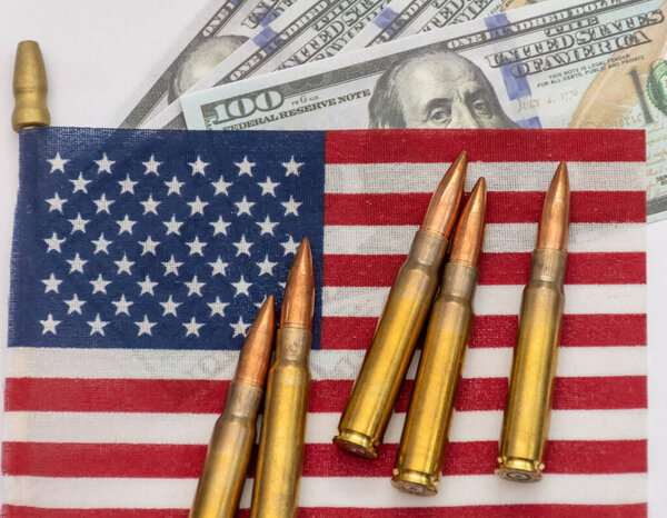 Several rifle bullets placed on a small United States flag with hundred dollar banknotes, symbolizing power, finance, and conflict.