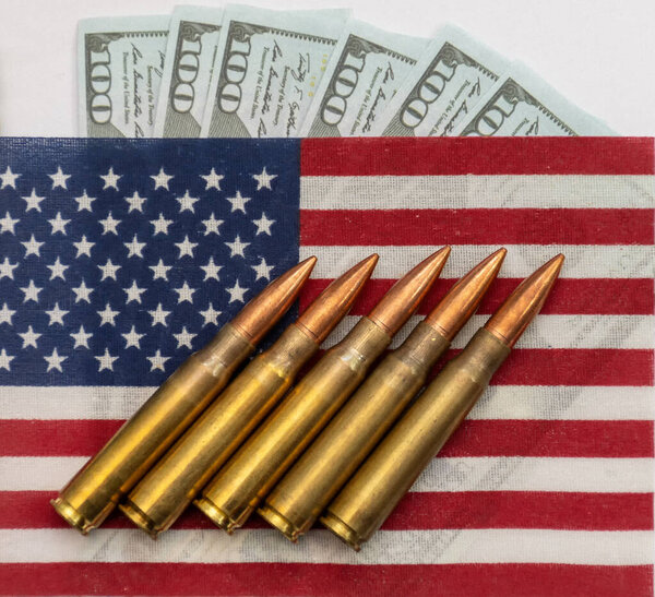 Several rifle bullets placed on a small United States flag with hundred dollar banknotes, symbolizing power, finance, and conflict.