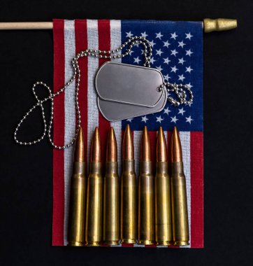 Siyah zemin üzerine dikey Amerikan bayrağına yerleştirilmiş gümüş künyeler ve hizalı tüfek fişekleri güç, hizmet ve vatanseverliği temsil ediyor..