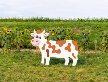 Parlak bir yaz gününde çiçek açan ayçiçeklerinin yanında çimenli bir tarlaya yerleştirilmiş kahverengi ve beyaz benekli neşeli ahşap inek figürü..