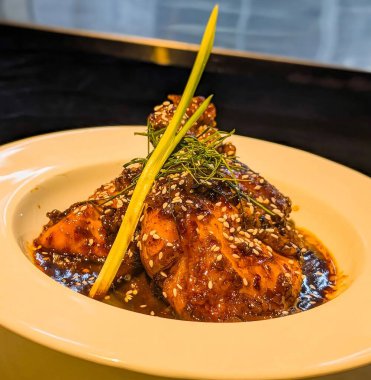 Close-up of tender glazed chicken pieces coated in a rich, glossy sauce and sprinkled with sesame seeds, elegantly garnished with fresh herbs in a white bowl