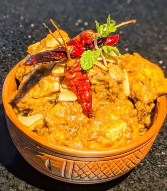 Close-up of a flavorful chicken curry topped with dried red chili, fresh herbs, and garlic slices, served in a rustic clay bowl.