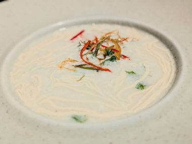 Close-up of a creamy coconut soup elegantly presented in a white bowl, topped with sliced red chili, fried shallots, and fresh herbs.