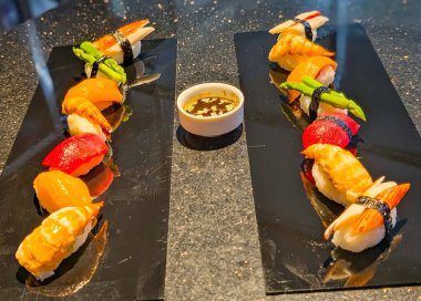 A colorful assortment of fresh nigiri sushi beautifully arranged on black plates with a bowl of soy sauce. The presentation highlights vibrant seafood, asparagus, and crab sticks.