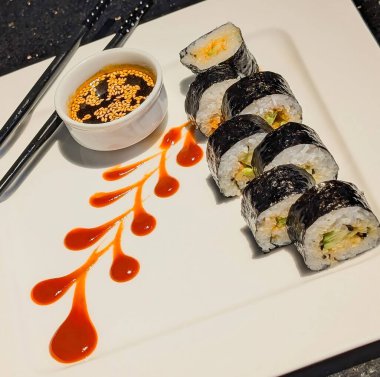 Fresh sushi rolls served with soy sauce and sesame seeds, elegantly plated with a drizzle of red sauce and crossed chopsticks.