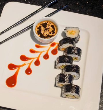 Fresh sushi rolls served with soy sauce and sesame seeds, elegantly plated with a drizzle of red sauce and crossed chopsticks.