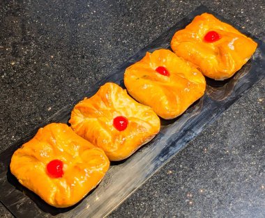 Four golden Danish pastries with glossy cherry topping arranged neatly on a black serving plate. Freshly baked dessert with flaky layers and rich custard filling.