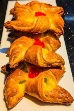 Close-up of freshly baked Danish pastries with golden flaky layers, creamy custard filling, and cherry topping on a white plate.