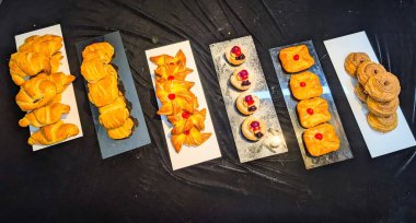Top view of assorted freshly baked pastries displayed on elegant serving trays. The collection includes croissants, puff pastries, and fruit danishes with glossy golden crusts and vibrant toppings.