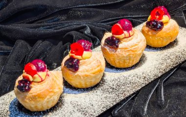 Close-up of freshly baked puff pastries topped with creamy custard, glazed cherries, and blueberry sauce.