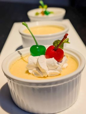 Description: Elegant vanilla custard served in a white ramekin, topped with whipped cream and colorful glazed cherries.