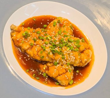 Close-up of crispy fried fish fillet served with rich soy garlic sauce, topped with sesame seeds and chopped spring onions.