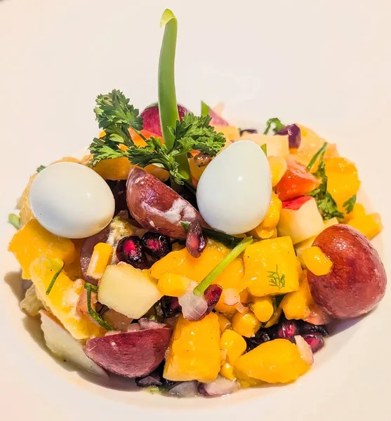 Colorful fruit salad with mango, grapes, pomegranate, corn, and herbs, topped with boiled quail eggs and garnished with parsley.