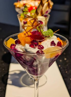 Beautifully arranged assorted fruit custard desserts served in elegant glasses, topped with cherries, wafer rolls, mint leaves, and vibrant mixed fruits.A delicious, colorful, and creamy sweet treat.