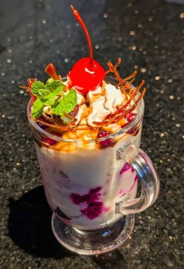 Close-up of a colorful layered fruit custard dessert served in a glass, topped with a bright red cherry, wafer rolls, mint leaves, and mixed fruits.