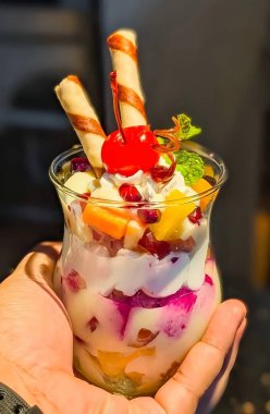 Close-up of a colorful layered fruit custard dessert served in a glass, topped with a bright red cherry, wafer rolls, mint leaves, and mixed fruits.
