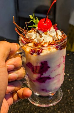 Close-up of a colorful layered fruit custard dessert served in a glass, topped with a bright red cherry, wafer rolls, mint leaves, and mixed fruits.