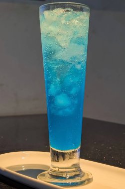 Chilled blue lagoon mocktail served in a tall glass filled with ice cubes on a white tray.The vibrant blue color and glossy texture create a refreshing,modern,and appetizing beverage presentation