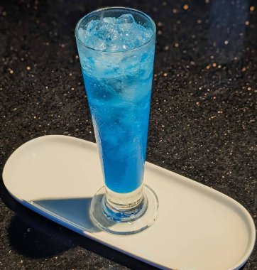 Chilled blue lagoon mocktail served in a tall glass filled with ice cubes on a white tray.The vibrant blue color and glossy texture create a refreshing,modern,and appetizing beverage presentation