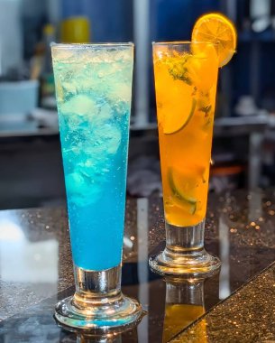 Two tall glasses of colorful summer mocktails,one bright blue lagoon and one orange citrus drink with lemon slices served on a glossy table.