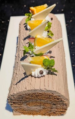 Elegant chocolate Swiss roll cake decorated with whipped cream, orange slices, white chocolate, and mint leaves. Beautiful dessert presentation on a white plate, perfect for bakery.
