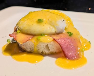 Closeup of a classic eggs benedict served on toasted bread with ham, poached egg, and creamy hollandaise sauce. A rich and savory breakfast dish beautifully plated and garnished with green onions.