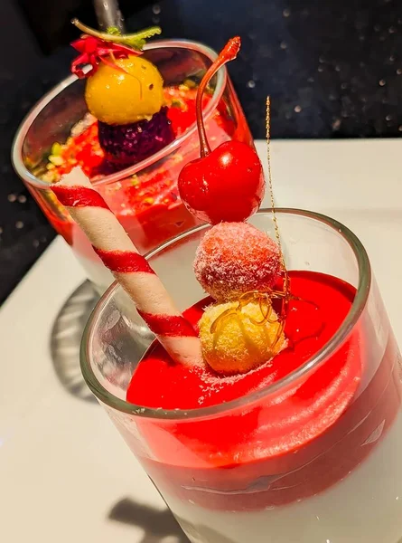 Two elegant individual desserts with a creamy white bottom layer and a shiny red berry sauce topping, garnished with fresh mint, a chocolate stick, and sweet sprinkles.