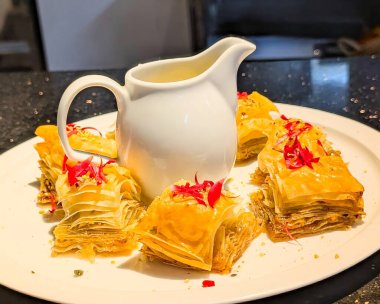 Close-up of golden, flaky baklava pieces arranged around a white jug of syrup, garnished with flower petals and crushed nuts.