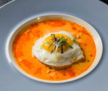 A gourmet dish featuring a perfectly fried egg served over a spicy, creamy broth, garnished with scallions, sesame seeds, and microgreens.
