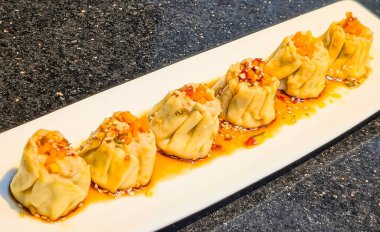 A row of steamed dumplings topped with diced vegetables and sesame seeds, served in a rich chili oil sauce.