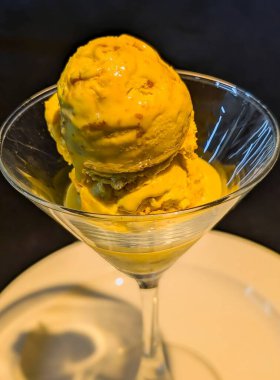 Canlı mango dondurması, görünür meyve parçaları ve parlak şurup, beyaz bir tabakta şeffaf bir martini bardağında servis edilir..