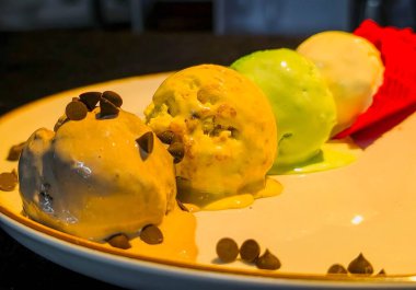 Parlak sarı bir tabakta kırmızı, yeşil ve kahverengi waffle külahlarıyla servis edilen canlı dondurma kepçeleri. Yaz yemeği promosyonu, menü tasarımı ve reklamcılık için ideal tatlı konsepti..