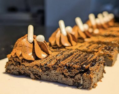 Çırpılmış çikolata kreması, kakao tozu ve zarif şeker incileriyle süslenmiş zengin çikolatalı brownilerin yakın çekimi, dokuyu ve hoşgörüyü vurgulamak için sıcak ışıkta fotoğraflandı..