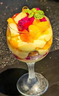 Berrak bir bardakta servis edilen renkli tropikal meyve tatlısı kremalı krema, kek parçaları ve ejderha meyvesi, kavun ve şeftali gibi çeşitli meyvelerle kaplı.