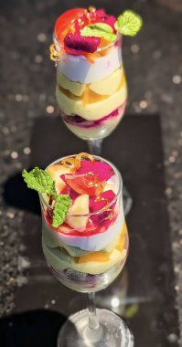 Berrak bir bardakta servis edilen renkli tropikal meyve tatlısı kremalı krema, kek parçaları ve ejderha meyvesi, kavun ve şeftali gibi çeşitli meyvelerle kaplı.