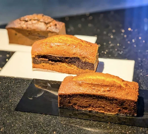 Ilık ışık altında parlak tabaklarda sergilenen zengin çikolata katmanlı altın mermer somun kekleri. Nemli doku ve zarif sunum onu fırın menüleri için ideal hale getiriyor..