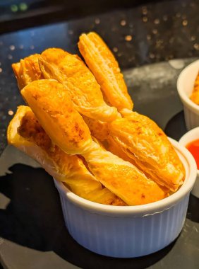 Koyu renkli bir tabakta, beyaz bir ramekin içinde servis edilen altın, pullu börek çubukları..